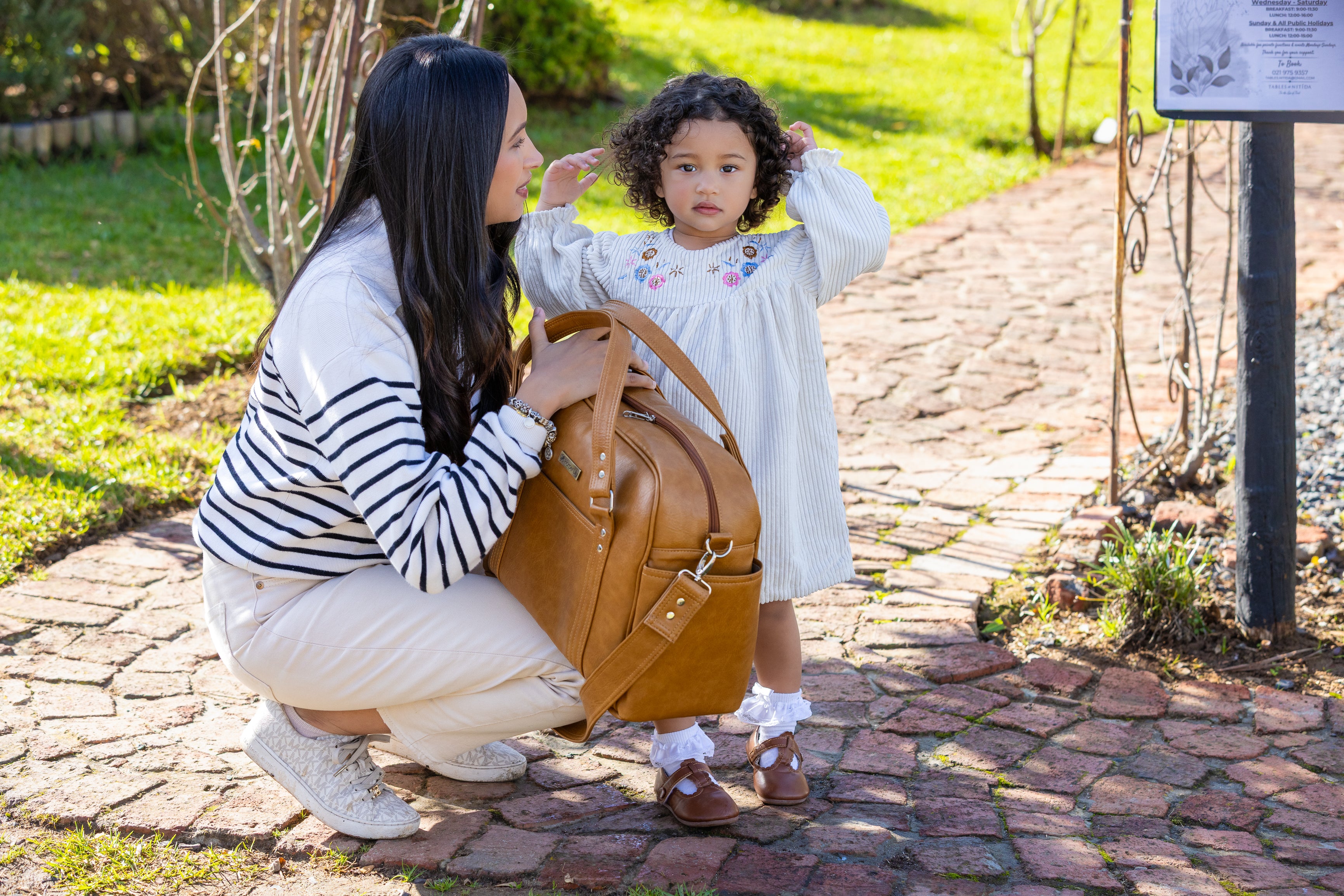 A woman and a young girl are outdoors on a stone path next to a tan leather shoulder bag. The woman is crouching beside the bag, and the girl is standing next to her in a white dress.